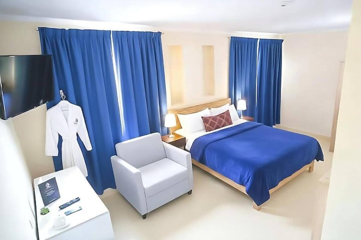 Une chambre d’hôtel avec un lit, un fauteuil blanc, une table de chevet avec une lampe et un téléviseur fixé au mur.