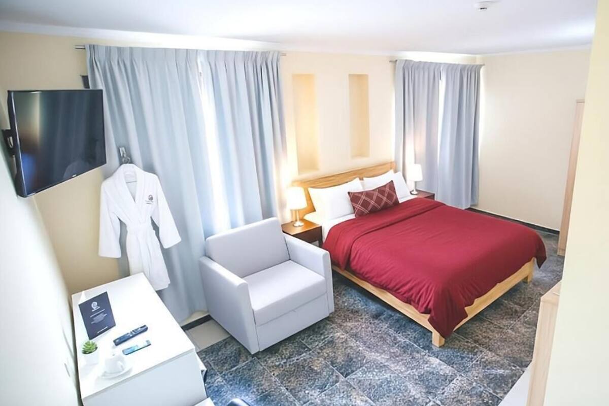 Une chambre d’hôtel avec un lit, un bureau avec un ordinateur portable, une chaise, un peignoir accroché au mur et un téléviseur fixé au-dessus du lit.
