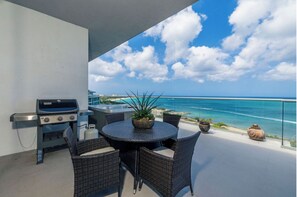 Un balcon avec un barbecue, des fauteuils en osier, une table ronde, des plantes en pot et une vue sur l’océan.