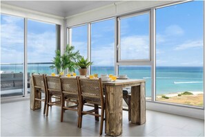 Un coin repas avec une table en bois et des chaises en osier, donnant sur une plage avec l’océan et un ciel dégagé.