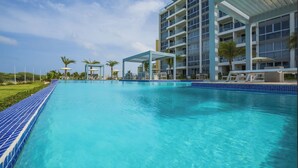 Una piscina con borde de azulejos azules, una zona de estar cubierta y un edificio de varios pisos con balcones al fondo.