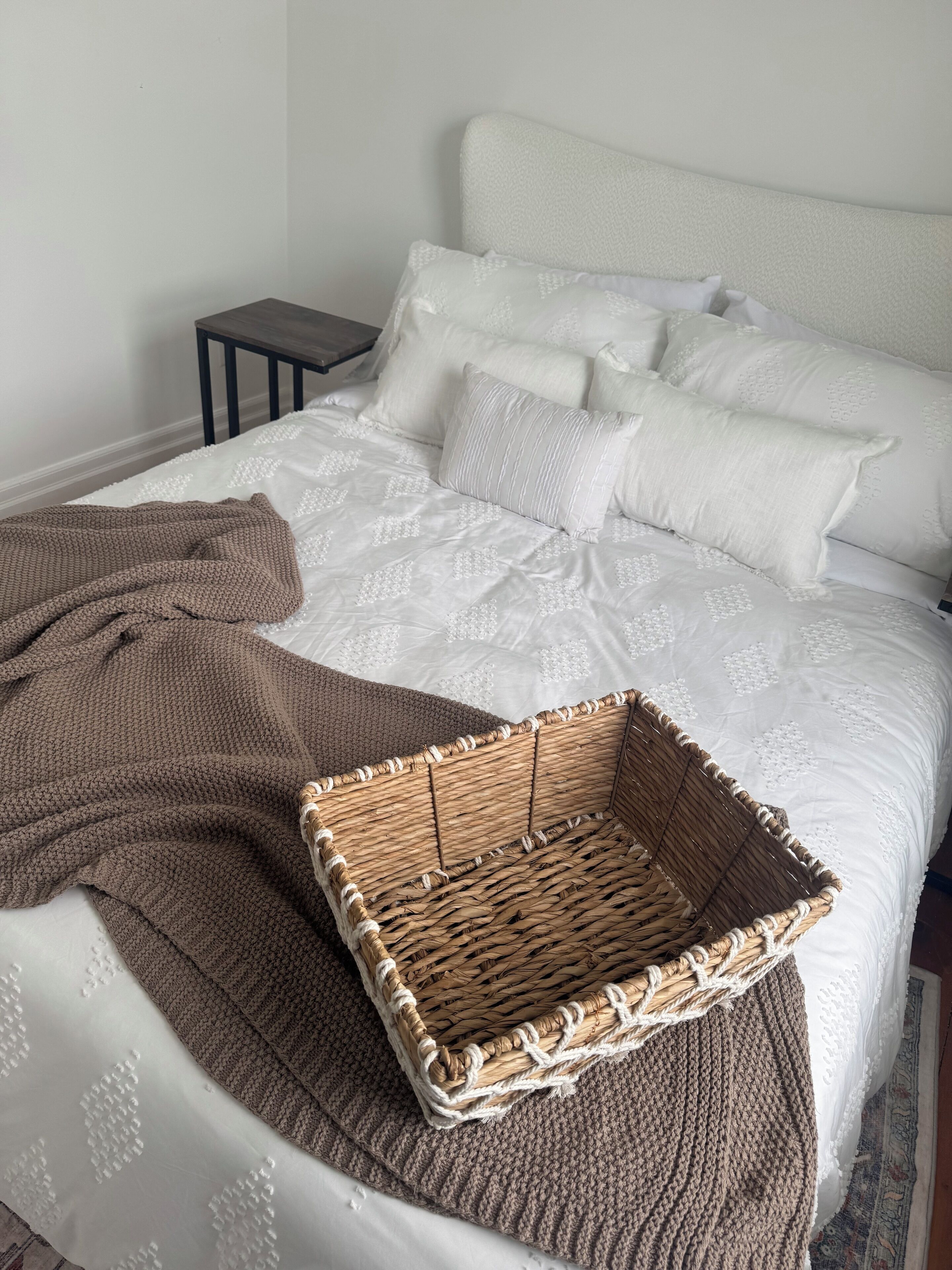 Un lit bien fait, avec une couette blanche et des oreillers, une couverture marron tricotée et un panier en osier posé sur le lit.