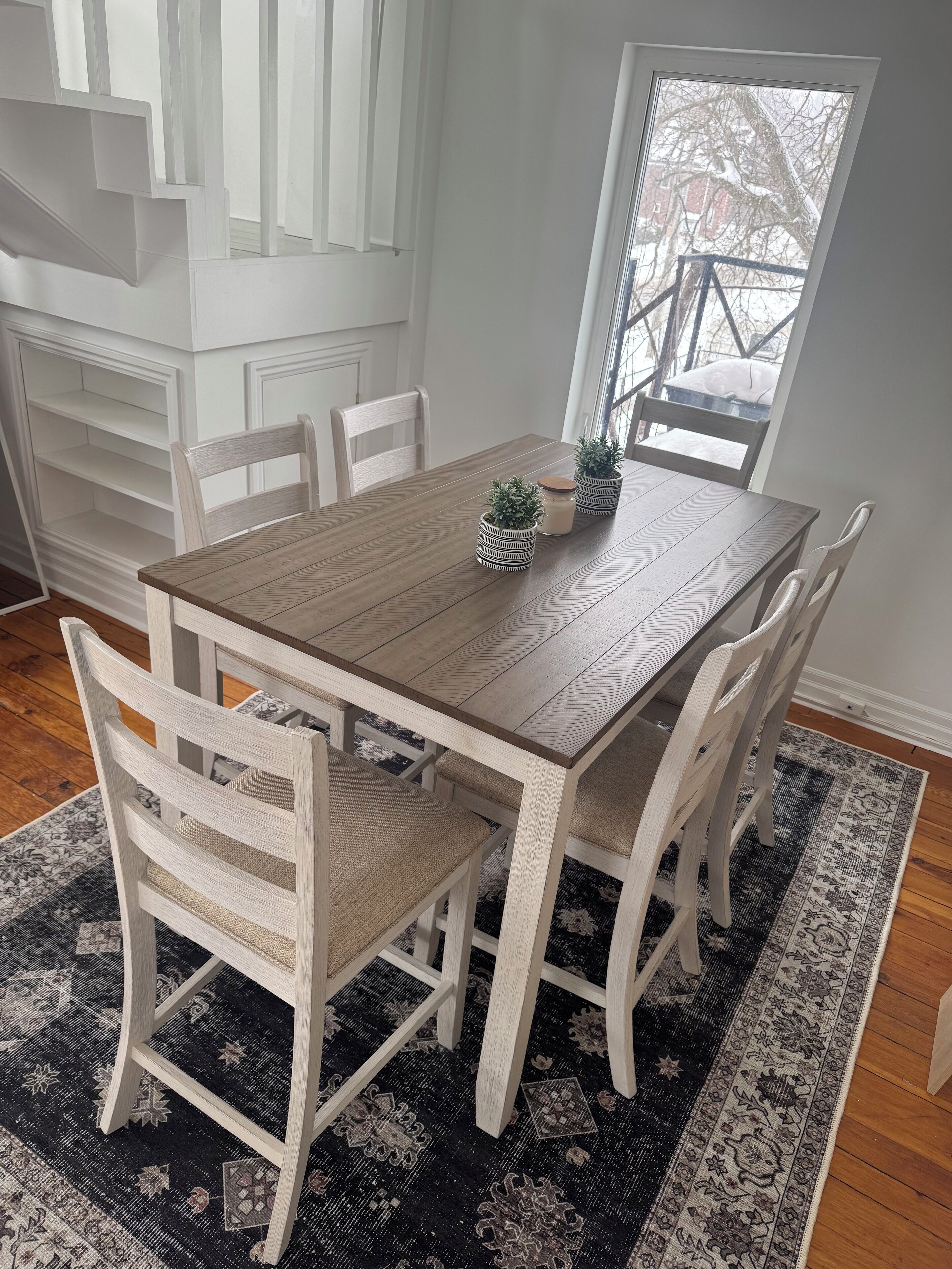 Un comedor con una mesa de madera, sillas blancas y un tapete estampado.