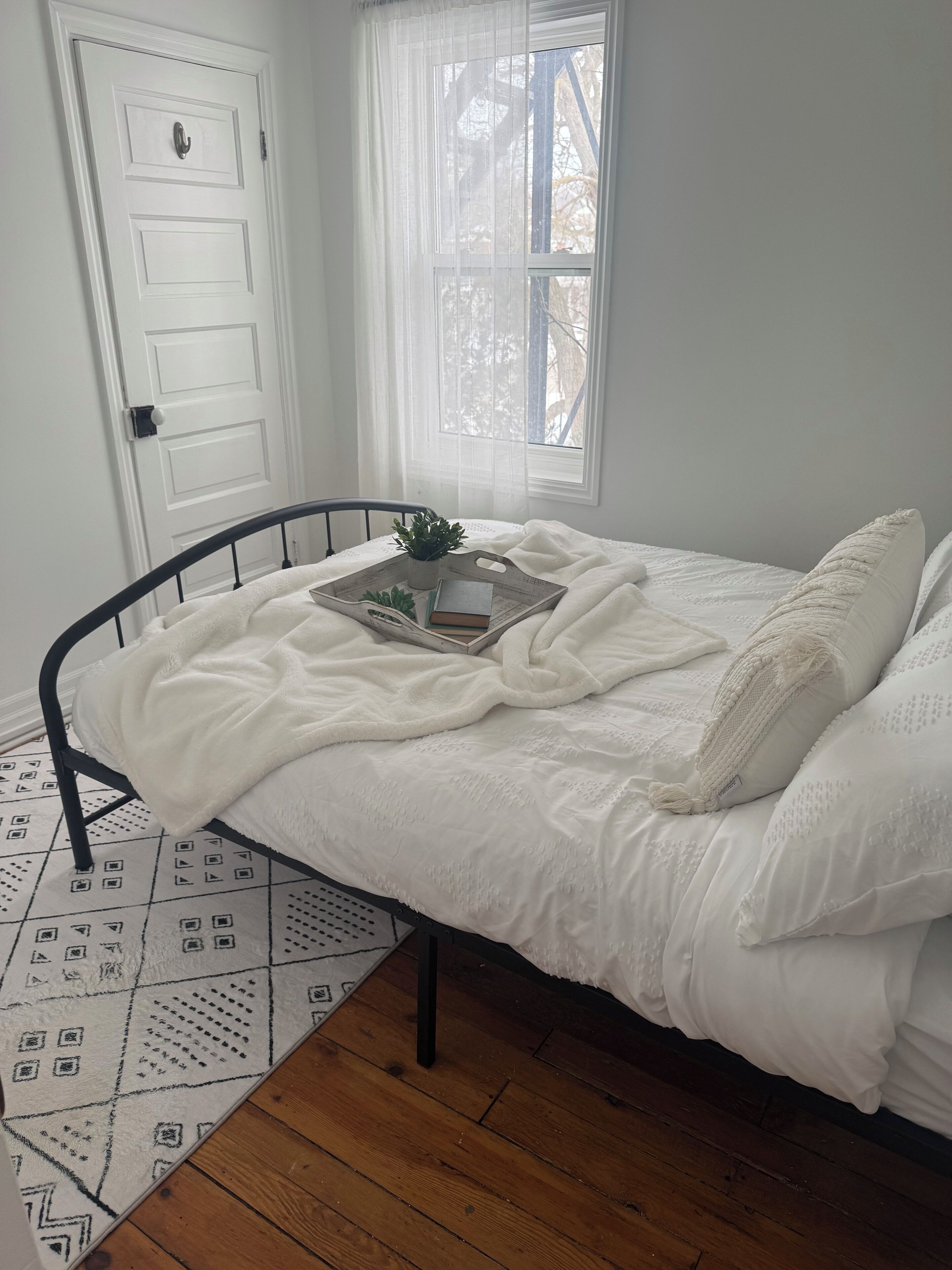 Een netjes opgemaakt bed met wit beddengoed, een dienblad met een plant en een boek, een raam met dunne gordijnen, en een witte deur.