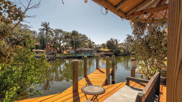 A wooden deck with a table and benches overlooking a calm body of water, surrounded by lush greenery and a few buildings in the background. - Canal Side Cocoa Beach 3BR 3BA Retreat (Cocoa Beach)
