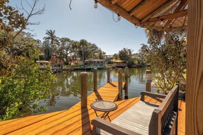 A wooden deck with a table and benches overlooking a calm body of water, surrounded by lush greenery and a few buildings in the background. - Canal Side Cocoa Beach 3BR 3BA Retreat (Cocoa Beach)