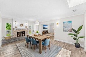 A spacious living room with a fireplace, a dining area with a wooden table and blue chairs, and a large potted plant.