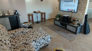 Olohuoneessa on kulmasohva, kiviseinälle asennettu televisio, pieni pöytä, jonka päällä on shakkilauta, ja kitara, joka nojaa seinään.
