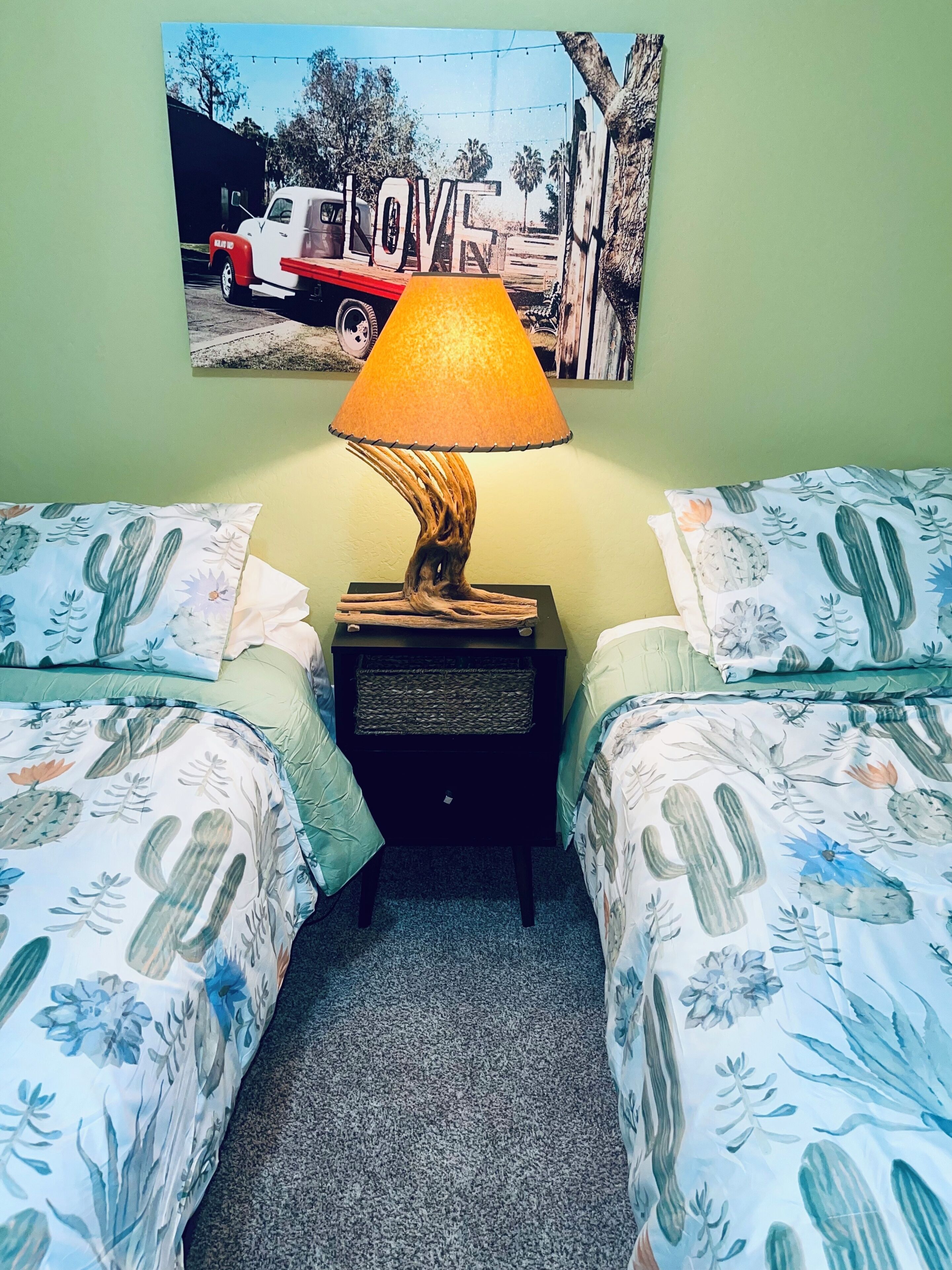 Una habitación con dos camas, una mesita de noche con lámpara y un cartel en la pared.