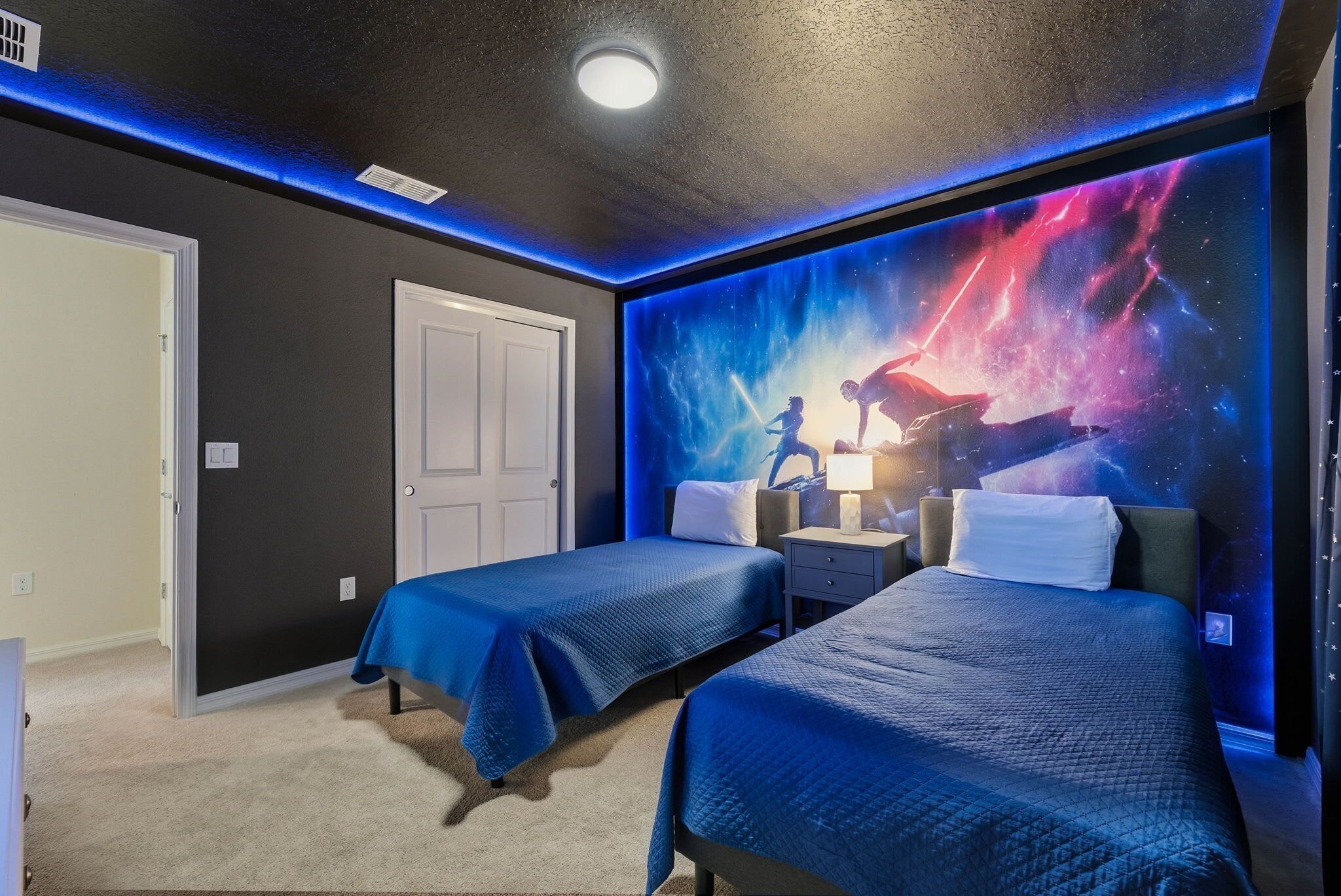 A hotel room with two beds, a nightstand, a wall mural of a cosmic scene, and a door to another room.