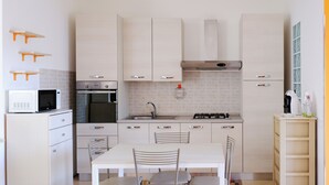 Eine kompakte Küche mit weißen Schränken, einer Mikrowelle, einem Backofen und einem Gasherd. Es gibt einen Esstisch mit Stühlen sowie eine kleine Regaleinheit mit Gegenständen darauf.