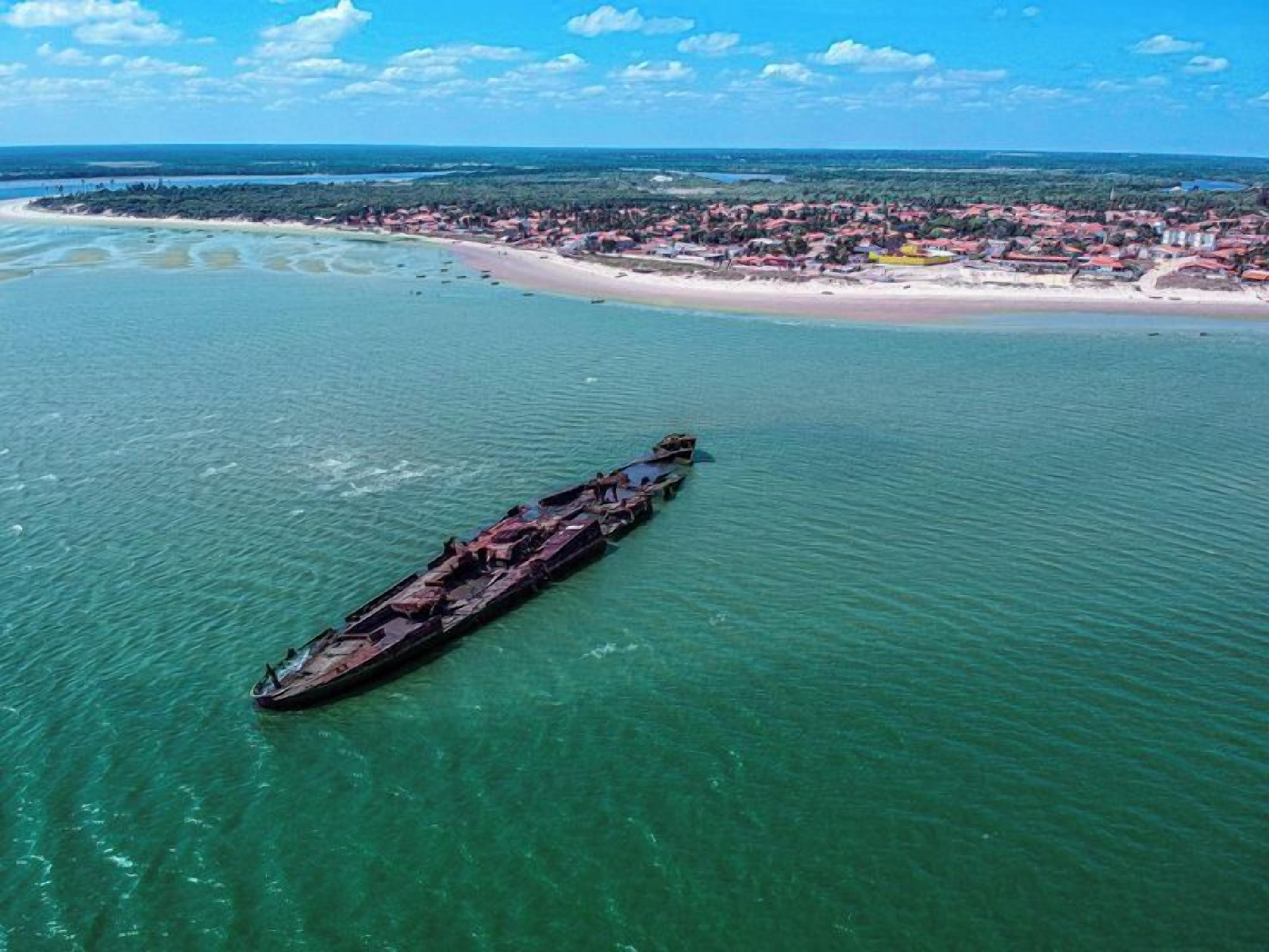 Vista aérea de una zona costera con un gran naufragio en el agua, una playa y un pueblo al fondo.