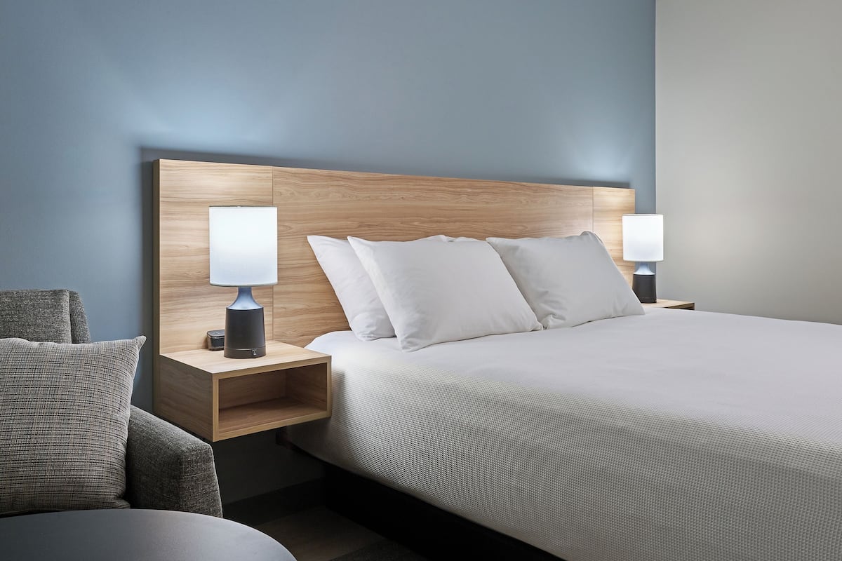 A modern hotel room with a wooden headboard, a large bed with white linens, two bedside tables with lamps, and a grey armchair.