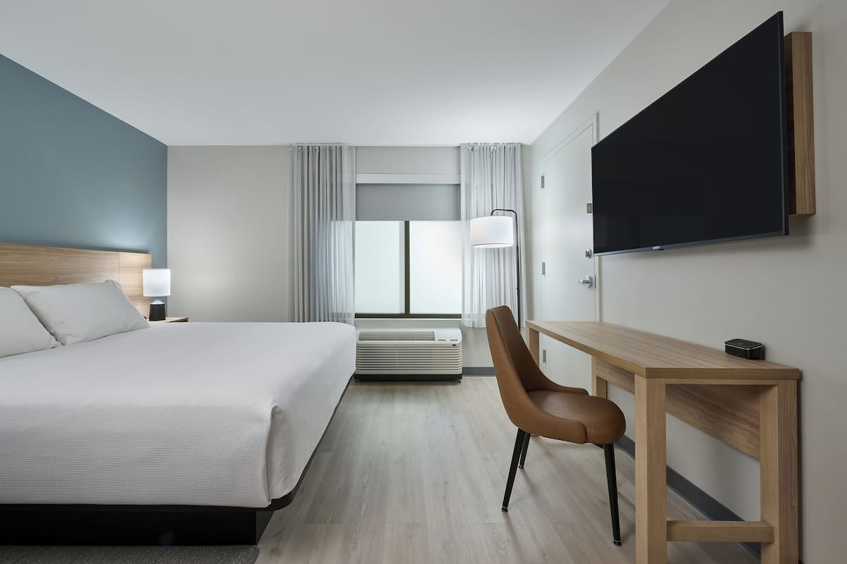 A modern hotel room with a large bed, a desk, a chair, a flat-screen TV, and a window with curtains.