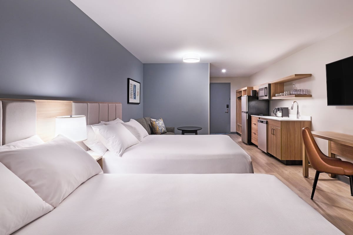 A hotel room with two beds, a desk, a chair, a TV, and a kitchenette.