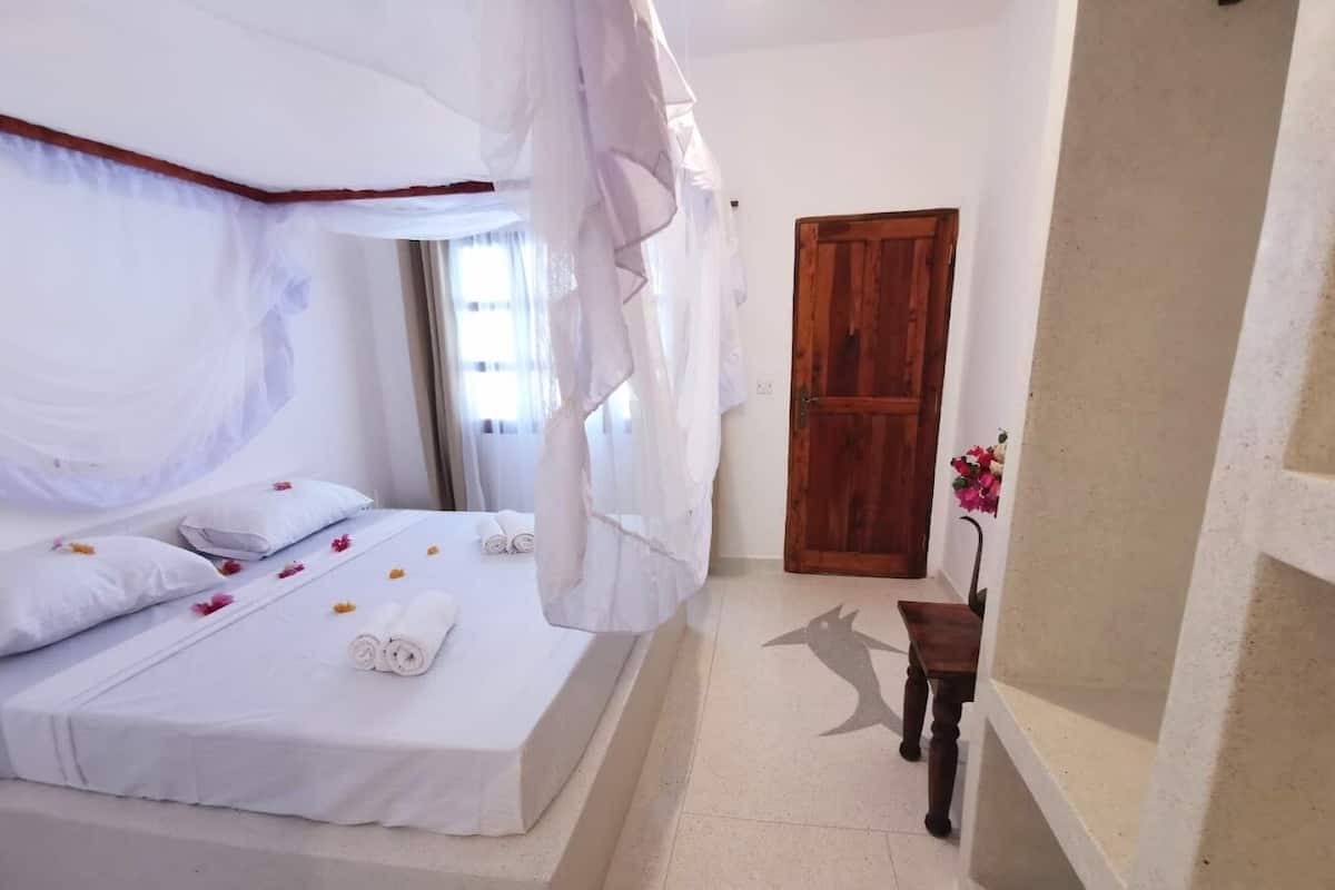 A bedroom with a bed, white curtains, a wooden door, and a small table with a vase.