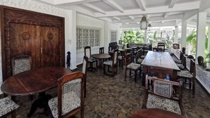 A spacious dining area with a large wooden table and multiple chairs, a decorative wooden door, and a view of lush greenery outside. - Thami House Watamu (Watamu)