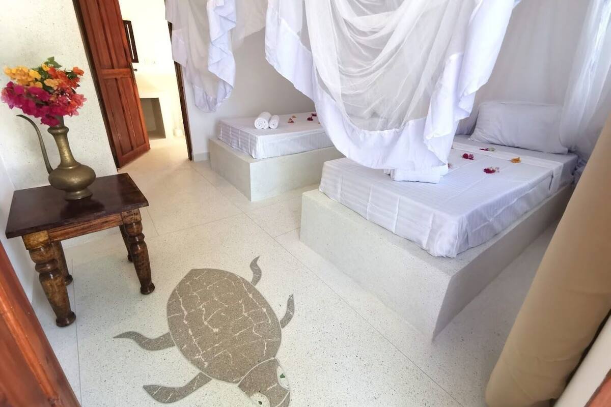 A bedroom with a bed under a white canopy, a bedside table with a vase of flowers, and a turtle mosaic on the floor.