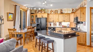 A modern kitchen with wooden cabinets, a central island with bar stools, a dining area with wooden chairs, and a large window with curtains. - Estes Park Retreat | 2 Kings | Near RMNP | ADA Friendly (Estes Park)