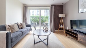 Un salon moderne avec un canapé gris, une table basse ronde, un téléviseur posé sur un meuble en bois et une grande fenêtre avec des rideaux.
