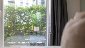 Une vue depuis une fenêtre, avec une table et un pot de fleurs à l’intérieur, un banc et de la verdure à l’extérieur.
