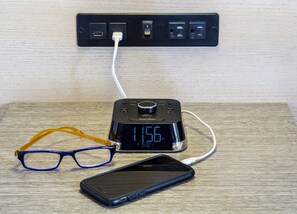 Una regleta con varios enchufes, un reloj digital que marca las 11:56 AM, un teléfono inteligente y un par de gafas sobre una superficie de madera.