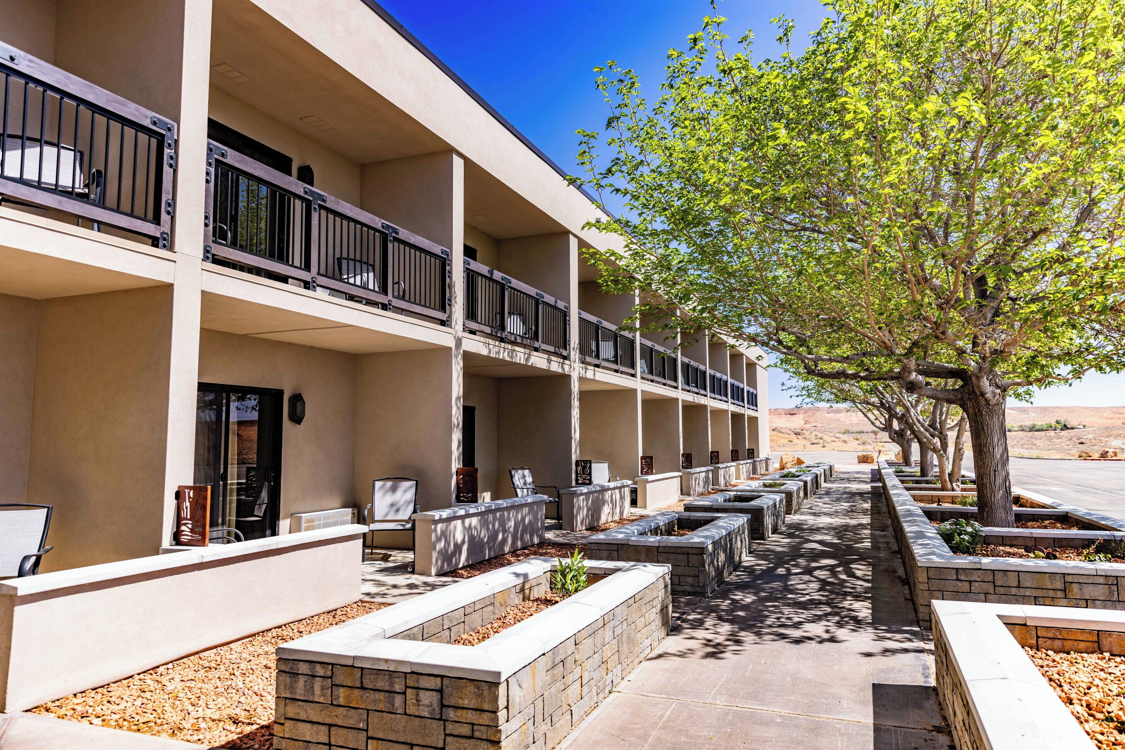 A multi-story hotel with outdoor seating, a tree-lined pathway, and a desert backdrop.