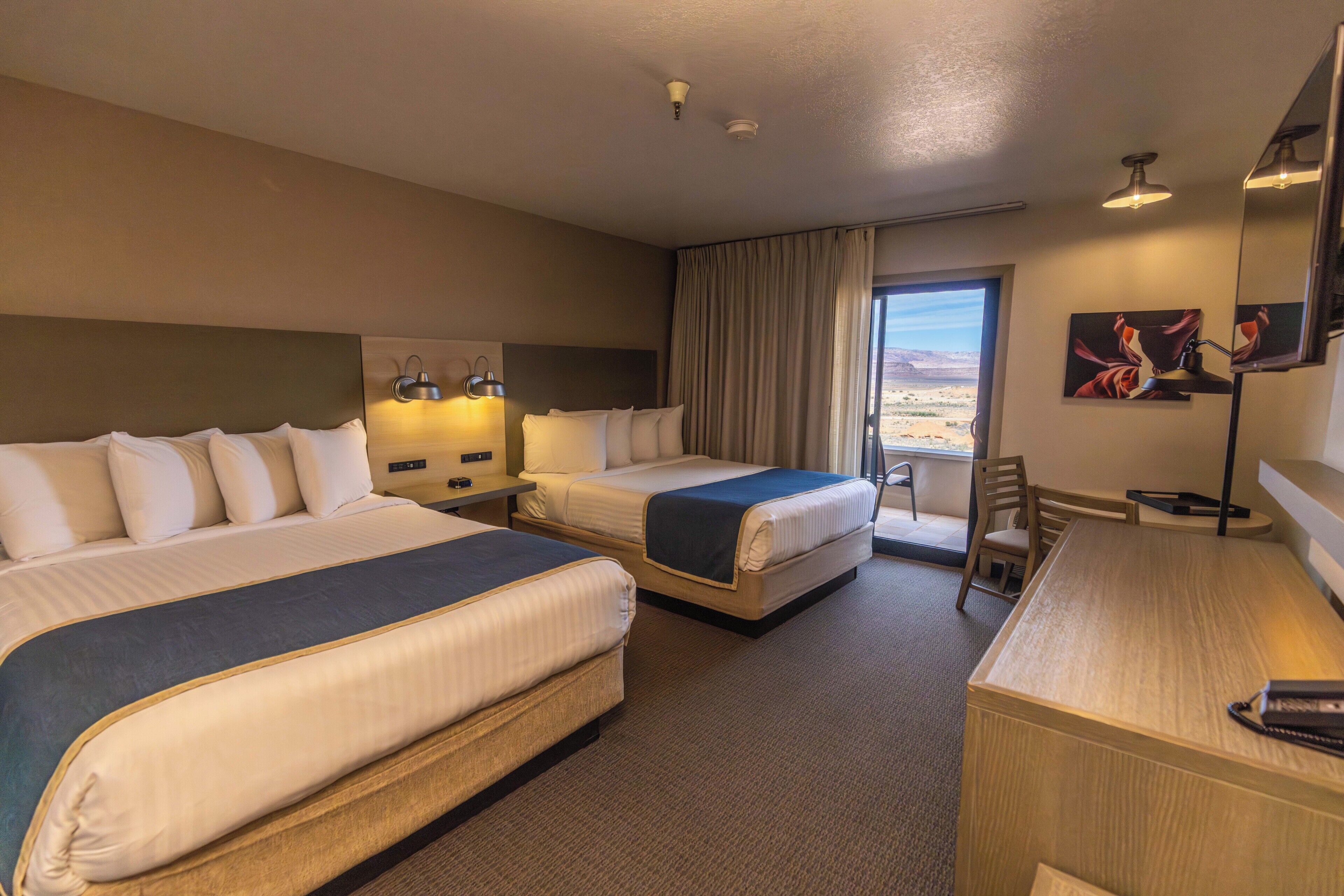 A hotel room with two beds, a desk, a chair, a large window with a view, and artwork on the wall.