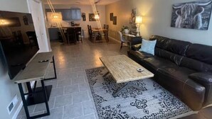 Een woonkamer met een bruine leren bank, een houten salontafel en een vloerkleed met dessin. Achterin is een eethoek met een tafel en stoelen.