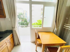 A small kitchen with a wooden table and chairs, a window with curtains, and a view of buildings and greenery outside. - Stay Central-Prestige Address &Private Comfort. (Thành phố Hồ Chí Minh)