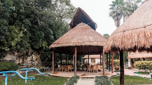 Un espace extérieur aménagé, abrité et doté de tables et de chaises, entouré d’un sentier de jardin et de constructions à toits de chaume.