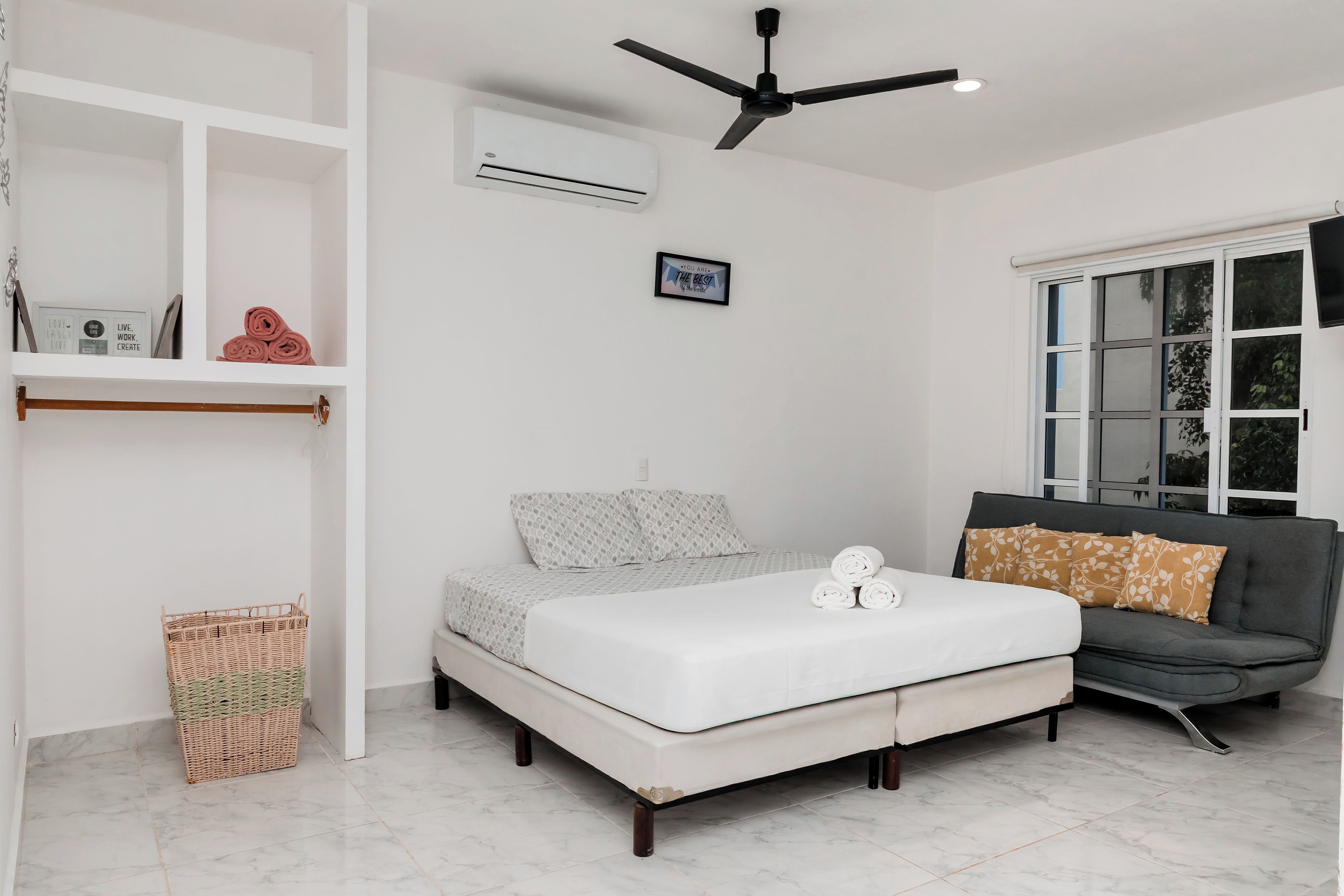 A bedroom with a bed, a grey sofa, a ceiling fan, an air conditioning unit, and a window with blinds.