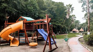 Une aire de jeux avec un toboggan et des balançoires, entourée d’arbres et bordée d’un sentier.