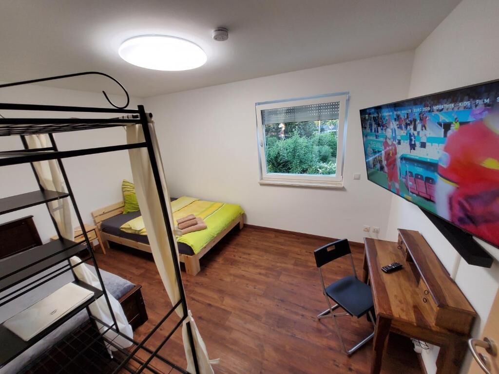 Een slaapzaal met stapelbedden, een bureau, een stoel, een televisie en een raam met uitzicht op groen.