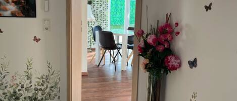 A room with a wooden floor, a door leading to a balcony, a wall clock, a framed picture, and a vase with flowers.