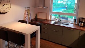 A modern kitchen with a large window, a dining area with a white table, and a clock on the wall. - Ferienwohnungen im Grund 22 (Hoppegarten)