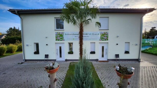 A building with a sign indicating it is a guesthouse. There are two potted plants in front of the entrance. - Ferienwohnungen im Grund 22 (Hoppegarten)
