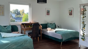 A room with two beds, a desk, a chair, a hanging lamp, and a window with a view of trees and buildings. - Ferienwohnungen im Grund 22 (Hoppegarten)