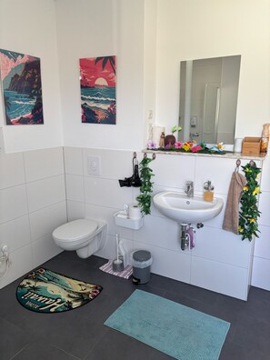 Un baño con inodoro, lavamanos y espejo. Se observan dos cuadros colgados en la pared, un perchero y un pequeño cubo de basura.