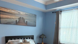 A bedroom with a bed, a nightstand, a lamp, a painting, and a window with curtains.