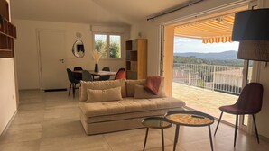 Een woonkamer met een bank, een eettafel en stoelen. Er is een balkon met uitzicht op het landschap.