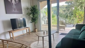 Een moderne woonkamer met een flatscreen televisie, een houten bankje, een ronde tafel met glazen, een groene bank en een grote kamerplant.