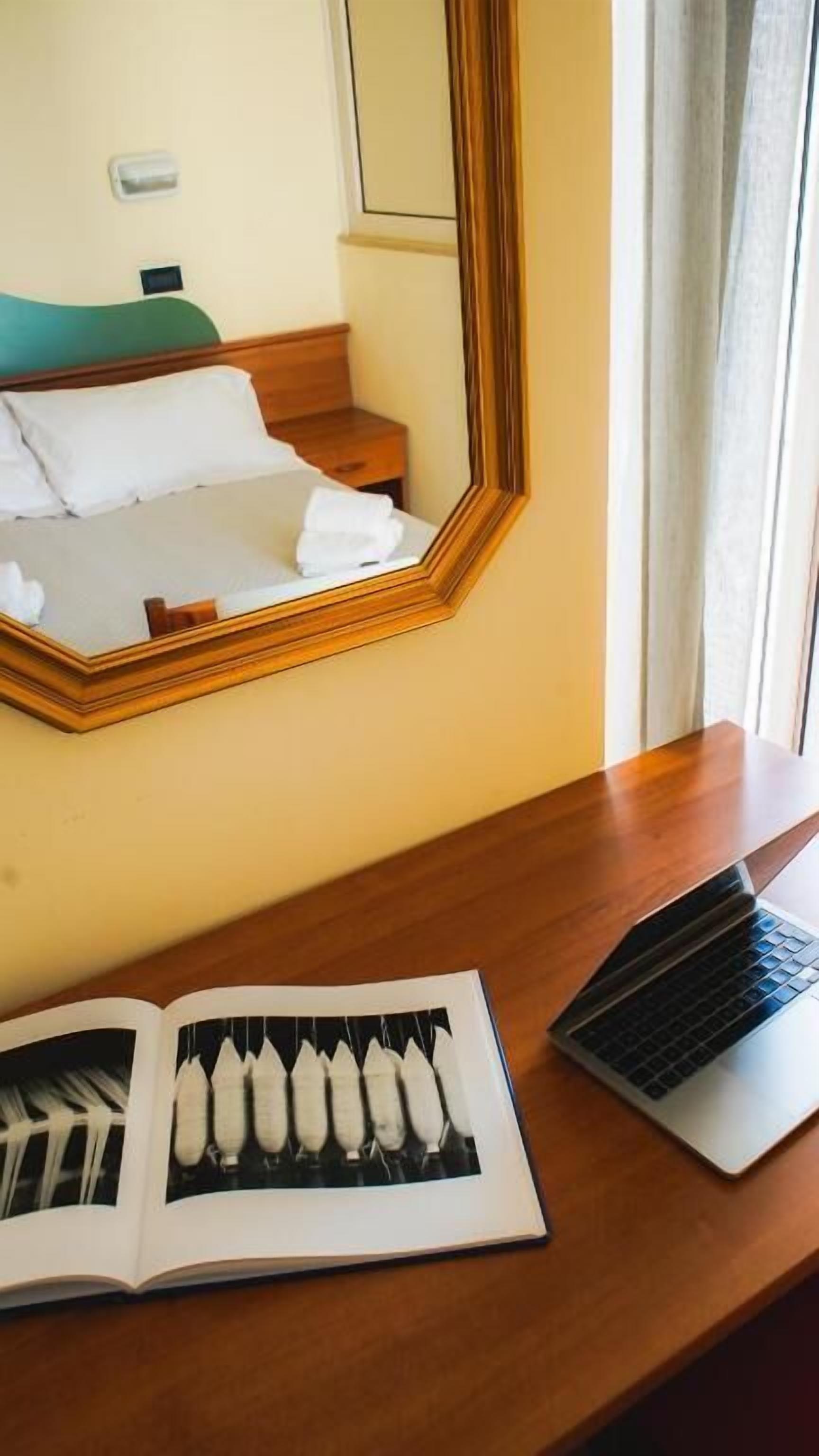 Quarto de hotel com uma cama, mesa de cabeceira, um laptop e um livro aberto.