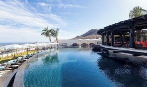 Une piscine avec des chaises longues et des parasols, un espace de détente couvert et une vue sur l’océan et les montagnes.