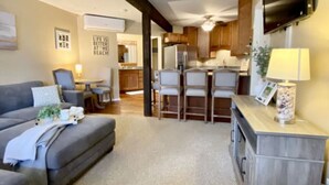 A modern living room with a grey sofa, a small table with a lamp, and a kitchen area with wooden cabinets and a TV mounted on the wall. - Deerpath Retreat at Abbey Springs (Fontana-on-Geneva Lake)