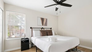 Ein Schlafzimmer mit einem Bett, einem Deckenventilator, einem Fenster mit Jalousien, einem Nachttisch und einem Teppich.