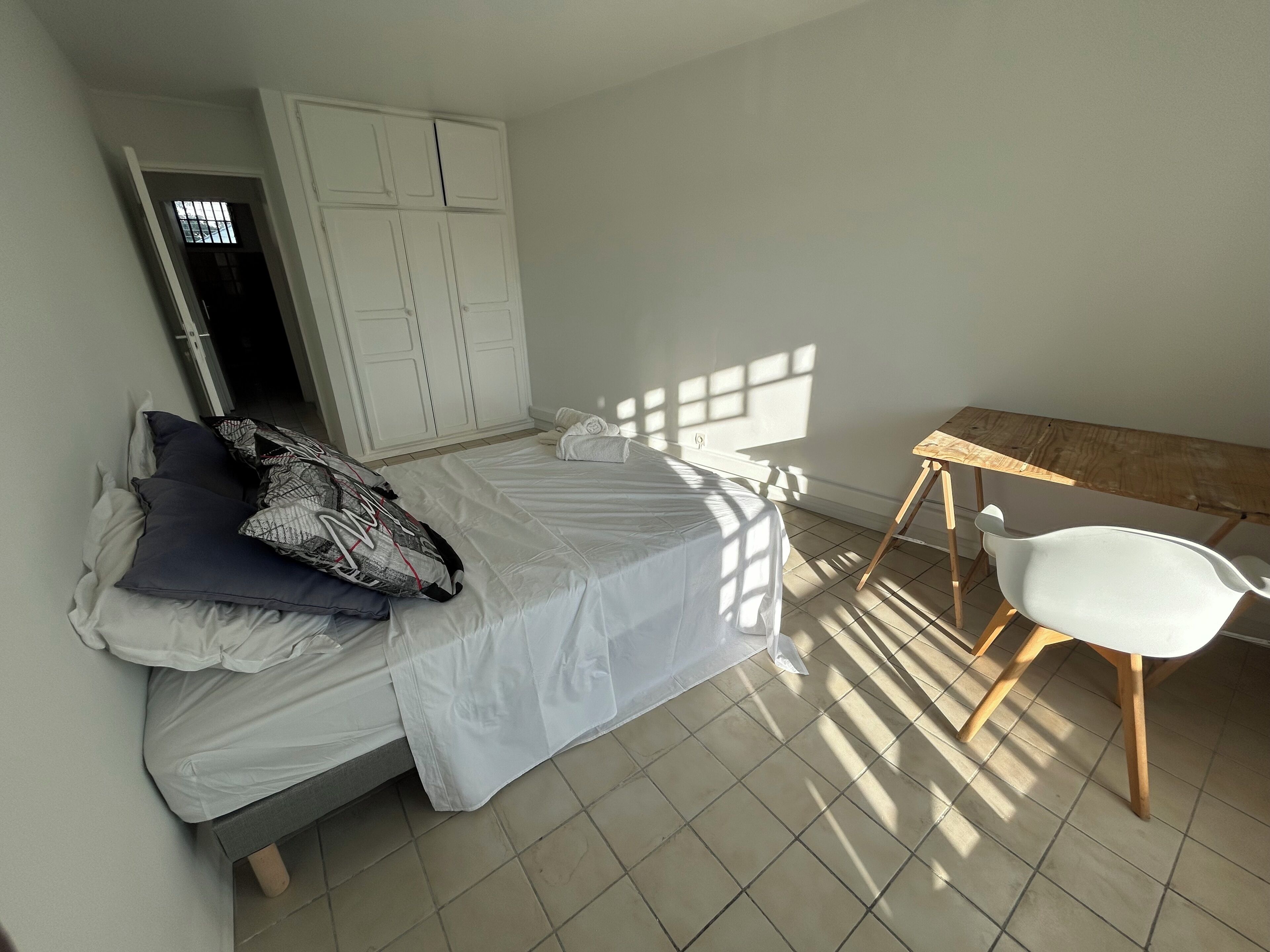A bedroom with a bed, a wooden chair, a wardrobe, and a window casting shadows.