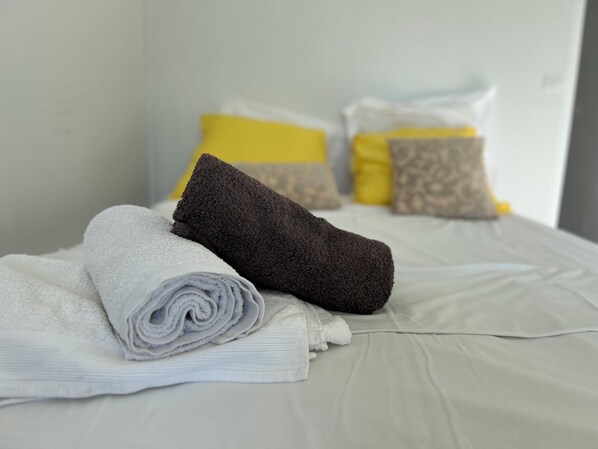 A neatly made bed with rolled white and dark brown towels, a white pillow, and two yellow pillows with a patterned design.