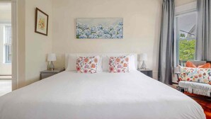 A hotel room with a large bed, two bedside tables with lamps, a painting on the wall, and a view of greenery outside. - Classic charm with modern comforts near Harvard. (Cambridge)