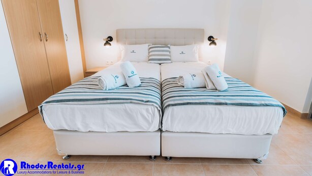 A double bed with striped bedding and two pillows, a nightstand with a lamp, and a wardrobe. - Rhodes Port Haven Suite (Rhodes)
